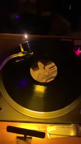The video shows a vinyl record spinning on a turntable. The record label has a distorted, striped pattern. A tonearm with a needle is positioned on the record. The turntable is lit by a warm, possibly yellow light. The environment appears dimly lit. A white object that looks like a USB drive lies near the turntable. The camera angle is from above, looking down at the turntable.