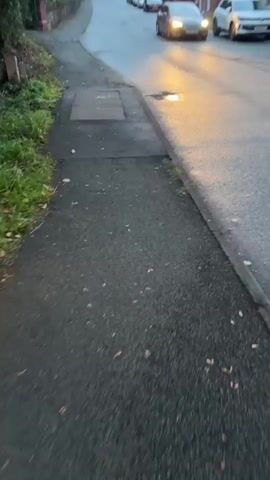 The video shows a sidewalk next to a street with cars driving by. The sky is overcast and it appears to be evening. There are houses and trees along the side of the road. A brown sign is partially visible in the upper left corner of the frame. A metal grate is set into the sidewalk.