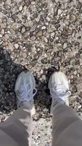 The video starts with a top-down view of a person's feet wearing white sneakers and khaki pants, standing on a pebbled surface. The person takes a few steps forward. The camera then shifts to an eye-level shot over a green railing, revealing a coastal town perched on cliffs overlooking the ocean. The town consists of closely packed buildings, with white and neutral colors dominating the architecture. The ocean is a vibrant turquoise color, and the sky is slightly overcast. The camera angle is positioned high up, giving a panoramic view of the landscape. In the foreground, below the railing, is a parking area with several cars.