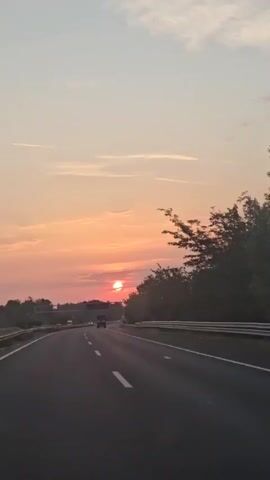 A time-lapse video shows a sunset over a highway. The sun is visible as a bright orange orb low in the sky, casting a warm glow. The sky is a gradient of pink, orange, and pale blue, with thin clouds scattered across it. Trees line the right side of the highway, and a few cars and trucks are driving on the road. A bridge or overpass is visible in the distance. The camera is mounted in a moving vehicle, giving a first-person perspective of the road ahead.