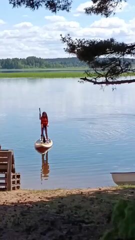 A person is paddleboarding on a calm body of water. The person is wearing an orange life vest and is holding a paddle. The paddleboard is a light brown color and is reflected in the water. In the foreground, there is a wooden bench and a sandy area with some grass. In the background, there is a line of trees and a blue sky with white clouds. The sun is shining.
