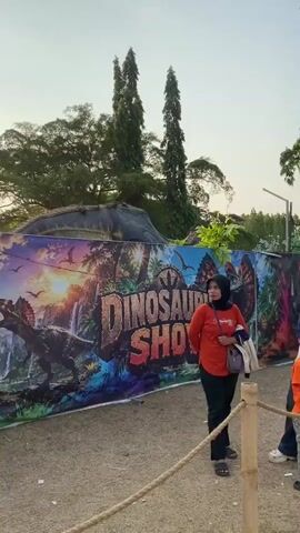 The video shows an outdoor scene with several people, mostly children and adults, standing near a colorful mural that depicts dinosaurs and the words "DINOSAUR SHOW." The mural is on a large banner. There are tall trees in the background and a bright sky, suggesting it is daytime, possibly late afternoon. Several children are interacting with each other and with adults. One child in an orange shirt and red hat is holding a rope. Another child in a green shirt is sitting on the ground. Adults are observing the children and the surroundings. There is a barrier made of ropes and wooden posts in the foreground. In the background, there is a structure that appears to be part of the dinosaur exhibit, with a dinosaur skeleton image on it. The overall atmosphere seems relaxed and festive, likely a public event or attraction.