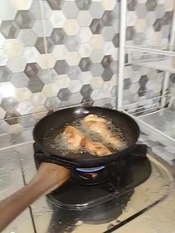 The video shows chicken legs being fried in a pan on a gas stove. The pan is black and has a wooden handle. The chicken legs are golden brown and appear to be nearly cooked. The oil in the pan is bubbling and sizzling. The stove is black and has a blue flame visible. The background shows a tiled wall with a hexagonal pattern and a white shelf unit with some items on it. A wooden spatula is seen on the left side of the pan, seemingly being used to adjust the chicken. The overall scene is a typical kitchen environment where cooking is taking place.