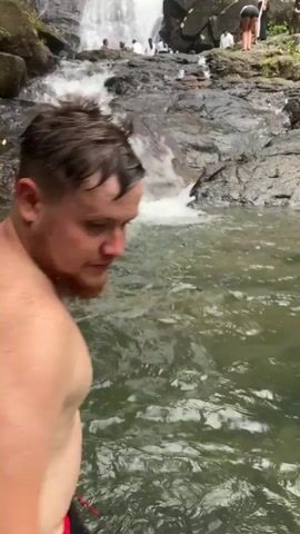 The video shows a man standing in a body of water, looking up at a waterfall. The waterfall cascades down a rocky cliff face surrounded by lush green vegetation. Several people are gathered on the rocks at the base of the waterfall, some sitting and others standing. The water is clear, and the flow of the waterfall appears strong.