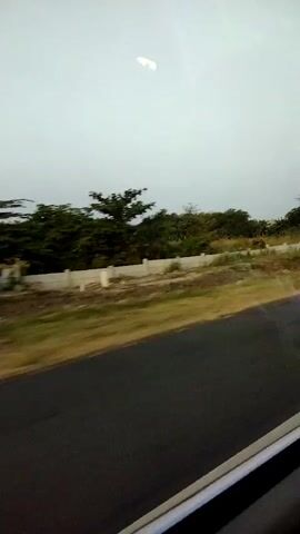 This is a still image taken from a moving vehicle, likely a car or bus, as evidenced by the motion blur in the foreground. The view is from a window, looking outwards. The scene is predominantly natural, with lush green vegetation on the left and center, forming a dense treeline. In the middle ground, there's a low, light-colored wall or barrier, possibly a retaining wall or safety barrier, with evenly spaced white cylindrical posts along its top. Beyond the wall, the vegetation continues. The foreground consists of a dark asphalt road, heavily blurred due to the vehicle's movement. The sky above is overcast and a pale gray, suggesting either early morning, late evening, or a cloudy day. There are no people or animals visible in the frame. The overall impression is of a roadside view during travel.