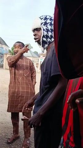 The video shows four dark-skinned men in what appears to be an altercation.  The main man in the foreground wears a dark t-shirt and an unusual winter hat with a white fuzzy brim and dark blue patterned sides.  He gestures aggressively with his right arm and has a scowl on his face.  In the background to the left, a man in a brown plaid outfit points to something off-screen.  Another man in a green shirt stands to the left as well. To the right, a man wearing a cap and a black and red striped jacket is looking at the main man, and blocking much of the shot. There is some indistinct background scenery of buildings and power lines, so the interaction appears to be outside.  Voices can be heard speaking in a language other than English.