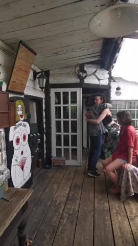 The video opens with a wide shot of an outdoor living space with a wooden floor and a white canopy overhead. There are two women on the left, one wearing a striped shirt and dark pants, the other in a red shirt and shorts. They are hugging each other, with the woman in the striped shirt briefly lifting the woman in red off the ground. A third woman in a black tank top and patterned skirt stands nearby, smiling. As the two women separate, the camera pans right, revealing a large body of water surrounded by rocky cliffs in the background. The outdoor space also features a wooden table with a watermelon and other items, a patterned sofa, and a brown armchair. The woman in the black tank top is now closer to the camera, smiling and looking off-screen. The woman in the striped shirt walks past the table, and the woman in red sits down on the sofa. The woman in the black tank top then walks past the sofa, and the video ends with a shot of the outdoor space and the water.
