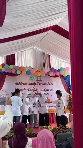 This video captures a stage event with a backdrop featuring balloons and text that appears to be in Indonesian. A group of young people, some wearing hijabs, are on stage. One young person, wearing a white t-shirt and a hijab, is holding a microphone and appears to be speaking or singing, while another young person, also in a white t-shirt, is standing next to them. A boy in a white t-shirt with text on it is facing the group on stage, holding a microphone. Another boy in a white t-shirt stands behind him. The event seems to be an "Akhirussanah" (graduation) and "Pentas Seni & Wisuda Tahfidz Al-Qur'an" (Art Performance and Graduation of Quran Hafiz). The audience members in the foreground are also wearing hijabs. The stage is covered by a tent with burgundy and white draping.