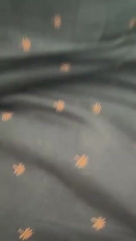 The video begins with a close-up of a dark fabric with orange dots. It then transitions to a shot focusing on the lower body of a person, showing their legs and knees. The person appears to be lying down on a bed, with dark blue bedding visible. In the background, a dresser is cluttered with various items, including bottles, what appears to be a witch's hat, and stuffed animals. The lighting suggests an indoor environment, possibly a bedroom. The person's feet are also visible, one resting on the bedding.