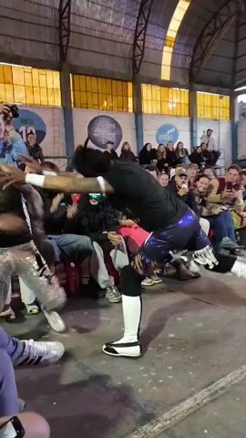 The video shows a wrestling match in a ring surrounded by spectators seated on chairs. Two wrestlers are actively engaged in the match, with one dressed in silver and the other in black and white attire with dreadlocks. The wrestler in silver falls to his knees, and the other wrestler delivers a chop to the back of his head. Spectators, some holding phones, react and observe the event. The environment appears to be an indoor arena with yellow-tinted windows. Some spectators are smiling or taking photos, showing their engagement with the wrestling match.
