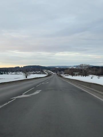 The video shows a first-person perspective from a vehicle traveling on a two-lane asphalt road through a winter or early spring landscape. The road, which appears damp or wet in sections, stretches straight ahead and then curves gently to the right in the distance, descending and ascending with the terrain. White dashed lines separate the lanes, and solid white lines mark the shoulders.

On both sides of the road, wide expanses of snow-covered ground are visible, suggesting fields or open areas. Scattered bare-branched deciduous trees stand out against the white snow, with one prominent tree alone in the field to the left of the road. Beyond these fields, dense dark green evergreen forests cover rolling hills and distant mountains, creating a rugged horizon.

The sky is overcast with a mix of light grey and white clouds. A faint strip of yellowish-orange light is visible near the horizon line, partially obscured by the clouds, suggesting either early morning or late afternoon. The overall atmosphere is serene and somewhat muted due to the cloudy conditions and winter scenery. The sound of driving is audible, indicating the vehicle is in motion. No specific location or text is discernible.