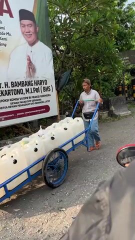 A man is pushing a cart with multiple large white water containers down a dirt road. He is wearing a light-colored shirt, blue pants, and a brown hat. The cart has blue metal framing and two wheels. Behind the man is a large poster with a picture of a smiling man in traditional clothing. The background shows green trees and foliage. The scene appears to be outdoors in a rural or semi-rural setting. The camera is positioned low and to the right, possibly from the perspective of someone on a motorcycle, with a portion of a red vehicle visible in the foreground.
