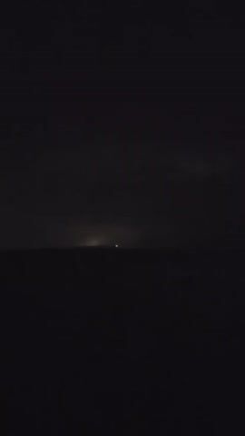 The video shows a dark scene with faint lights on the horizon, likely a distant city or ships. The sky is dark and cloudy, with occasional flashes of lightning illuminating the clouds. The ocean is calm and dark. The video is shaky, suggesting it was taken from a moving platform, possibly a boat.