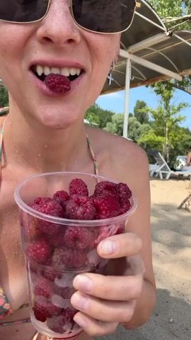 The video shows a close-up of a person holding a clear plastic cup filled with fresh raspberries. The person is wearing sunglasses and a bikini. In one shot, the person is seen putting a raspberry into their mouth. The background suggests a beach scene with sand, trees, and a body of water where other people are visible swimming and relaxing. The overall atmosphere appears to be casual and summery.