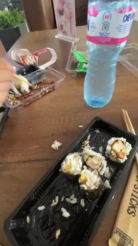 A person is using chopsticks to eat sushi. There is a bottle of water on the table, along with empty sushi containers and some leftover rice. The person is wearing a black t-shirt. The environment appears to be a restaurant or dining area.