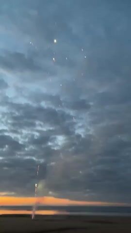 A time-lapse video shows fireworks exploding in the sky over a beach and ocean at dusk. The sky is filled with clouds, and the sun is setting, casting an orange glow on the horizon. Fireworks are seen exploding in various colors, including red, white, and yellow. Smoke from the fireworks rises into the sky. The beach is empty, and the ocean is calm.