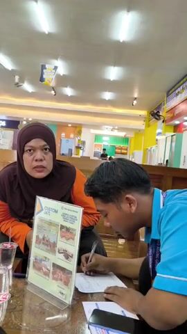 A woman wearing a maroon hijab and an orange shirt is seated on a wooden bench. In front of her is a table with two plastic cups and a menu stand. A man in a blue polo shirt with "YOUNG GROUP" written on it is seated opposite her, writing on a piece of paper. The background appears to be a retail store with shelves and displays. The lighting is fluorescent.
