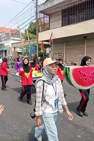 A group of people, mostly young women wearing hijabs and red shirts with yellow accents, are participating in a parade on a street. They are carrying a long, flat, rectangular object supported by poles. Some are waving. Other people are lining the street, watching the parade. The environment appears to be an urban setting with buildings and overhead wires. The weather is sunny.