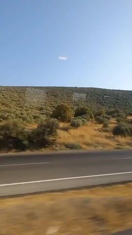 The video shows a view from a moving vehicle. The foreground features a highway with a white line marking its edge. Beyond the highway, a dry, grassy field stretches towards a sloping hill covered in vegetation, primarily olive trees. The sky is clear and blue, with a faint, linear arrangement of lights visible high in the distance. The overall impression is one of a sunny, rural landscape.