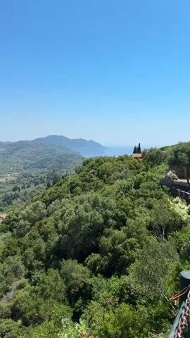 A still image captures a panoramic view of a lush green landscape under a clear blue sky. The foreground is dominated by a dense cluster of trees with dark green foliage. Beyond this, the terrain slopes downwards into a valley dotted with more trees, olive groves, and scattered houses. In the mid-ground, a winding road can be seen, and further in the distance, rolling hills stretch towards the horizon. The background reveals a vast expanse of the sea and more distant landmasses. A small red-roofed building is perched on a hilltop in the upper right portion of the frame. The overall impression is one of a serene, sun-drenched rural or mountainous region.