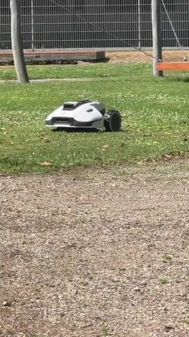 A robotic lawnmower is autonomously navigating a grassy area. The lawnmower is white and gray with a black wheel on the right side. It appears to be in motion, with its front facing slightly to the left. The setting is outdoors on a sunny day, suggested by the bright lighting and shadows. The grass is green and dotted with small white flowers. In the background, a chain-link fence and wooden structures are visible, hinting at a park or recreational area. A gravel path occupies the foreground, creating a textural contrast with the grass. There are no people visible in the frame. The overall scene is peaceful and suggests a well-maintained outdoor space.