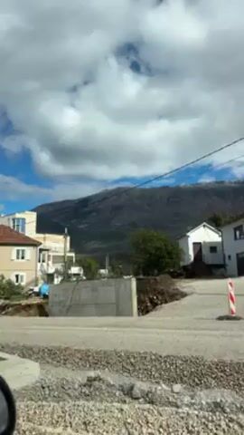 The video shows a view from a car driving on a dirt road through a rural area. A partially constructed concrete structure is visible beside the road, with a mountain range in the background. Several houses are visible to the left and right of the road. The sky is partly cloudy.