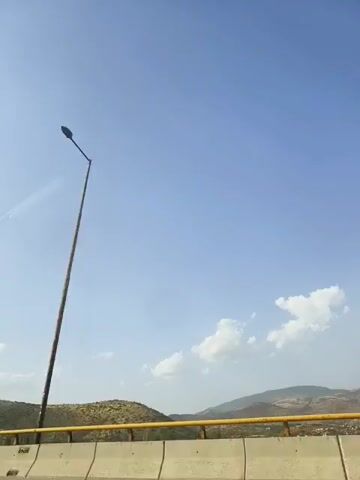 The video shows a view of a clear blue sky with a few white clouds. In the foreground, there is a concrete barrier and a yellow railing. Beyond the railing, there are hills with sparse vegetation. A tall lamppost is visible on the right side of the frame. The camera appears to be moving.