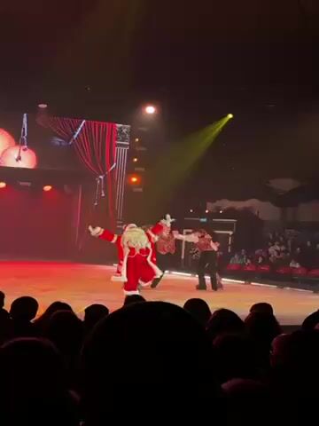 A brightly lit stage is the setting for a Christmas-themed ice skating performance. In the foreground is a partially visible audience. On the ice, Santa Claus is prominently featured, interacting with other skaters in festive attire. The skaters perform lifts and spins, their movements dynamic and energetic. The background includes theatrical elements like red curtains and decorative columns, as well as large, stylized lanterns hanging above the stage. The lighting shifts between yellow and red, enhancing the overall festive atmosphere.