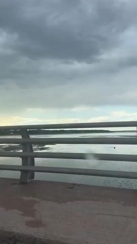 The video shows a view from inside a vehicle, looking out at a body of water under a cloudy sky. The vehicle is moving, as evidenced by the shifting scenery. In the foreground, there is a concrete road and a metal guardrail. In the middle ground, there is a body of water with some structures or buoys visible. In the background, there is a distant shoreline with trees and buildings. There is also a blue sign with a white question mark and the number 150m. The overall atmosphere is overcast and potentially stormy. There are no people or animals clearly visible in the video.
