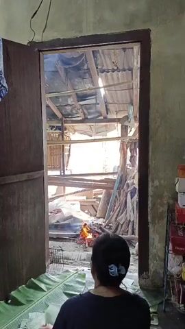 A video shows a person facing away from the camera, seated on large green leaves spread on the floor. They are looking through an open doorway into an outdoor area. A fire burns in the outdoor area, with smoke rising. The outdoor area appears to be a makeshift structure with a corrugated metal roof, wooden beams, and piles of wood and materials. The person has dark hair tied back with a clip. The indoor area appears to be a simple room with a textured wall.