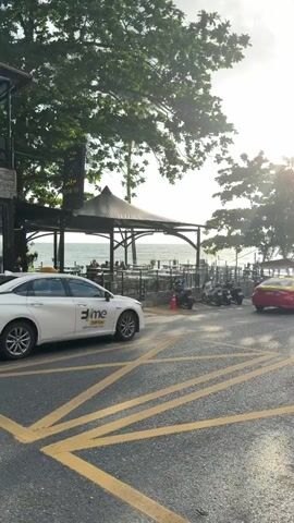 The video shows a street scene near a beach with several vehicles and trees. In the foreground, there are yellow lines painted on the asphalt, indicating a parking or driving lane. A white sedan with the logo "ime" on its side is parked on the left. Further down the street, a red and yellow taxi is visible. Several motorcycles are parked or moving around. The background reveals a body of water, likely the ocean, with trees lining the coastline. The sun is shining brightly, casting a glare and creating a hazy effect. The sky is partly cloudy, with some dramatic clouds visible. There are also signs and utility poles along the street. People can be seen in the distance, some near the vehicles and others possibly on the beach. The overall impression is a busy roadside environment with a coastal backdrop, likely during the daytime.