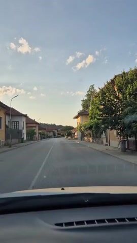 This is a first-person perspective video clip taken from a car driving down a street in Lesidren, Bulgaria. The scene is a quiet village street lined with houses and trees. The time of day appears to be late afternoon or early evening, indicated by the warm sunlight casting long shadows across the road and the soft lighting on the buildings. The sky is a clear blue with scattered white clouds, suggesting pleasant weather.

The subjects are the houses lining the street, the road itself, and the natural elements of trees and sky. The houses are of a traditional architectural style, with tiled roofs and visible windows. Some houses have gardens or yards with greenery. The road is paved and has a white dividing line. The overall impression is one of a peaceful, rural setting.

The activity taking place is simply driving through the village. There are no people or other vehicles visible in the immediate vicinity, contributing to the serene atmosphere.

Notable details include the specific time of day and weather conditions, which create a calm and inviting ambiance. The architecture of the houses and the presence of mature trees offer subtle cues about the location's character and potential age.

There is no visible text within the frame of the video.