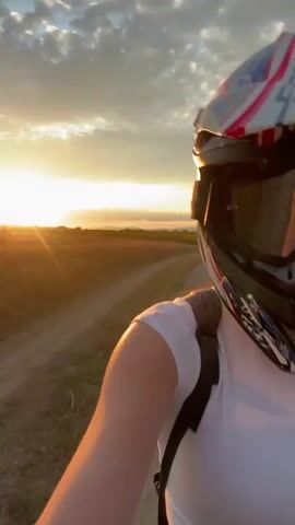 A person wearing a white shirt and a dirt bike helmet is in a field. The helmet is decorated with an American flag pattern and has large goggles. The person raises their hand, wearing a glove, and makes a peace sign gesture. The background shows a dirt path and dry grass, with a sunset visible in the distance. The camera is held close to the person, creating a selfie-like perspective.