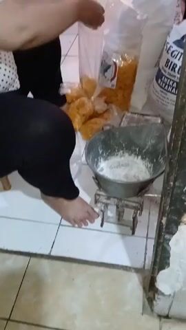 A person is in a room with tiled flooring. They are squatting on the floor wearing black pants and a white polka dot shirt. The person is holding a large plastic bag with a blue and white label, pouring yellow food items into a metal scale that has flour in it. There are several other plastic bags filled with the same yellow food items on the floor. A large white bag with Indonesian text is also visible in the background. The scene appears to be a domestic or commercial kitchen or storage area.