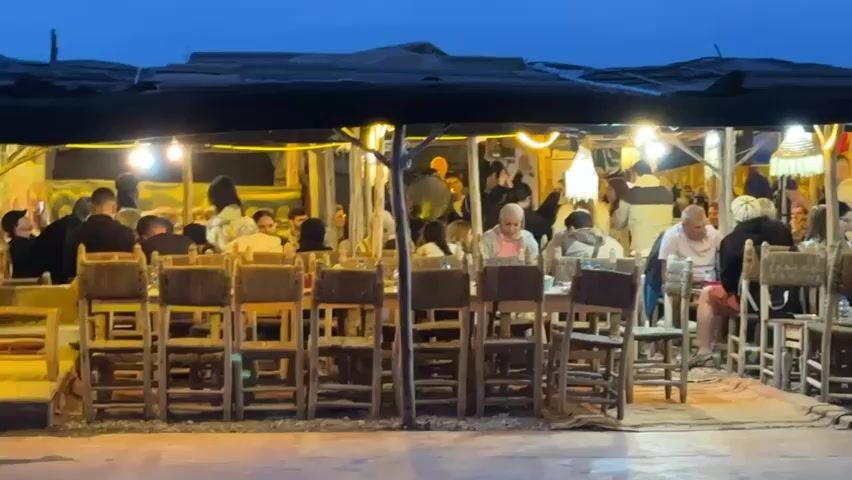 The video presents a lively outdoor gathering at dusk or night in Sidi Badhaj, Morocco. Numerous people, predominantly adults, are seen socializing and dining under a dark canopy illuminated by string lights and various overhead lamps, some featuring decorative woven shades. The sky in the background is a deep twilight blue.

The scene features multiple long, rustic wooden tables paired with tall, high-backed chairs constructed from wood and woven materials. Many individuals are seated, enjoying meals and conversations, while others are standing and mingling. The ground beneath the seating area appears to be sand or dirt, suggesting a desert or camp-like setting.

A festive atmosphere pervades the gathering, enhanced by music playing in the background. Several people are observed swaying, clapping, or moving to the rhythm, indicating dancing or active enjoyment of the music. The attendees generally appear relaxed, happy, and engaged in the social event. There are no visible signs of inclement weather. No text is discernible within the frame. The overall impression is one of a vibrant and enjoyable evening social event in a unique, traditionally inspired Moroccan outdoor venue.