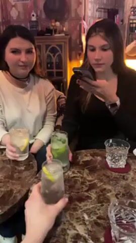 Two young women sit side by side at a marble-patterned bar or table. One is taking a photo of a cocktail glass held by a third person, out of view, while the other woman smiles and holds her cocktail glass. The drink in the woman's hand who is taking a photo has lime in it, while the other woman's has a lemon. The bar has a vintage, dim aesthetic, with patterned wallpaper. The woman taking the photo is wearing a black shirt, the woman smiling is wearing a light colored top. A cut glass tumbler sits to the right. Music plays.