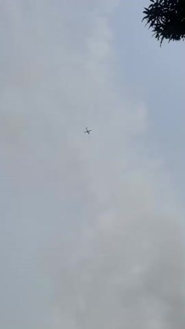 A drone camera shot from a low angle captures a cloudy sky with a small airplane visible in the distance. The plane is small in the frame and appears to be in flight. The top right of the frame shows the dark silhouette of tree branches. The video is 4 seconds long.