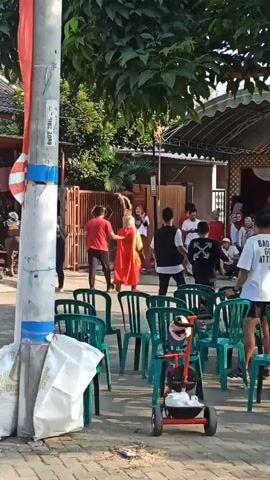 A group of people are gathered outdoors, possibly for an event, with many green plastic chairs arranged in rows. In the center of the frame, a person in a vibrant orange and gold costume with a headdress is standing and facing away from the camera. Another person in a red shirt is walking towards the costumed individual. The environment appears to be a residential area with trees and a building in the background. A utility pole is visible in the foreground on the left side of the image.