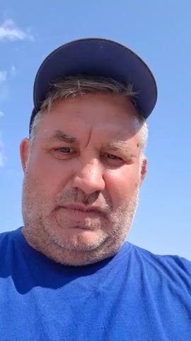 A man wearing a blue t-shirt and a blue baseball cap is filmed outdoors against a bright blue sky with a few wispy clouds. The man is looking directly at the camera and speaking. He has a round face, stubble, and appears to be middle-aged. The camera angle is close-up, focusing on his face and upper torso. The lighting suggests it is daytime.