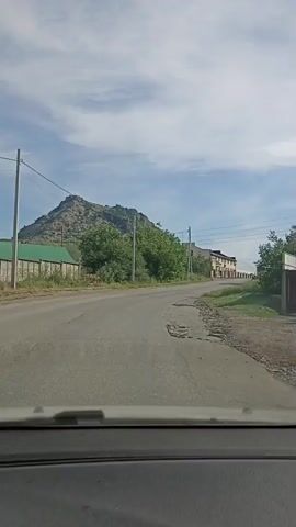 The video shows a view from inside a vehicle traveling on a road. The road is paved but shows signs of wear and tear, with a large pothole visible. On the left side of the road, there is a fence and some trees. In the background, there is a rocky mountain. On the right side of the road, there are buildings with light-colored walls and darker roofs, possibly a row of garages or storage units. The sky is partly cloudy. The video captures the movement of the vehicle as it drives along the road.