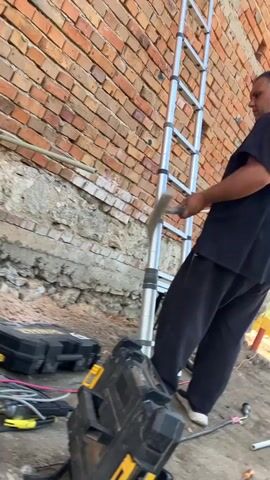 A man in a black shirt and black pants is holding a shovel and chipping away at the bottom of a brick wall.  A silver ladder is leaning against the wall.  There is a black tool box and a yellow tool box on the ground next to the wall. There is also a drill and some electrical cords on the ground. The man is right-handed. The ground is dirt and sand.  It is daytime and the sun is shining.