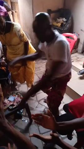 The video captures an indoor scene with several individuals. One person, wearing a white shirt and patterned shorts, is dancing. An older woman in a yellow dress dances beside him. There are several people sitting on the floor clapping along with the dancing. A tray with money and other items rests on a small table near the participants. The room appears cluttered, with a red chair, a trash bag, and miscellaneous objects visible in the background. The lighting is moderate, providing a clear view of the activities.