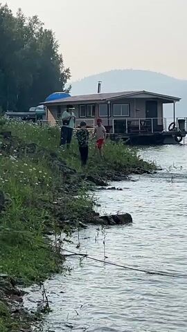Two children and an adult stand on the shore of a body of water, next to a floating house. The adult is wearing a hat and holding what appears to be a phone. One child is wearing a red hat and a white shirt with red shorts. The other child is wearing a patterned shirt and dark shorts. The child in the red hat appears to be throwing something into the water. The floating house has several windows and a railing. The shore is covered in grass and small white flowers. In the background, there are trees and hills. The water is rippling.