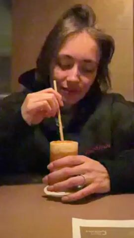 A woman is sitting at a table, drinking a beverage with a straw. She is wearing a black hooded sweatshirt and has a ring on her finger. The drink is an orange color and has a frothy top. There is a paper menu on the table. The background is a neutral wall.