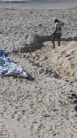 From an aerial perspective, the video depicts a sandy beach next to a body of water, presumably the sea, with an individual and some objects present.

An individual, clad in darker attire, is visible on the sand near a dark, amorphous spot. The person is in a position suggesting they are bending or crouching over something on the ground. Nearby, a pile of what appears to be crumpled or folded material, possibly fabric or tarp, lies on the sand. Additional indistinct objects are scattered to the upper right.