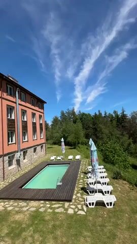 The video shows a bright sunny day at an outdoor resort. The first shot is of a rectangular turquoise swimming pool with a wooden deck beside a building with tan stucco walls. Several white lounge chairs and blue-and-white patio umbrellas surround the pool on a grassy lawn. A forest is visible in the background, and the sky is blue with wispy white clouds. The camera then pans upward and to the right, focusing on an outdoor patio furnished with a dark table and black chairs. The patio overlooks the forest and mountains. The sky remains a clear blue backdrop.