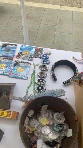 The video pans across a white-covered table displaying various items, likely at an outdoor market or stall. In the foreground left, there's a woven basket containing many small, round objects, some in clear plastic packaging. To the right of the basket is a small, decorative, dark-colored incense holder with a square opening, and next to it, a rectangular box containing what appears to be incense sticks. Behind the basket, a dark wooden box holds several small glass items, possibly shot glasses.

Moving up the table, there are several keychains with round metallic charms. To their right are multiple packages, each containing a circular metallic coin or medallion, some with blue and white packaging featuring images of landscapes or buildings. Further right, there's a display of colorful beaded necklaces and decorative hair accessories with intricate pearl and gem embellishments. A black headband adorned with similar embellishments is also visible. A light blue decorative fan lies partially open on the table.

In the middle ground, a white dog, possibly a poodle or bichon frise, is visible looking over the table. A rotating display stand holding numerous keychains with various charms is positioned behind the dog. Various flat, rectangular items, some in plastic sleeves, are spread across the table, depicting scenic views or artwork. A red Coca-Cola can with a straw sits among these items. A small, dark-colored wine bottle in packaging is also present.

In the background, people are walking by. One person, wearing brown pants and white shoes, is seen bending down near the table. Another person is partially visible in the upper left corner, with what looks like the wheel of a stroller. The environment appears to be an outdoor pedestrian area with paved ground and some greenery visible beyond the table. The lighting suggests it is daytime.