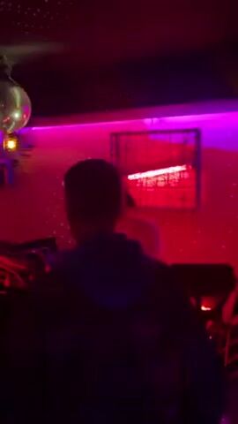 The video shows a person in the foreground, facing away from the camera, in a dimly lit, possibly nightclub, environment. In the background, another person is visible, seemingly operating DJ equipment. The room is decorated with red and purple lighting, including neon fixtures on the wall and a disco ball. The backdrop features a patterned surface.