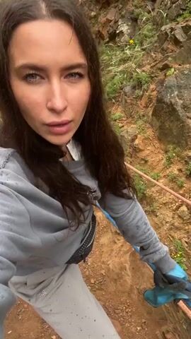 The video depicts a woman holding a camera while standing on a steep, rocky hillside. The hillside is covered in patches of green vegetation and reddish-brown soil. Several ropes are fixed to the hillside, leading down towards a sandy beach and ocean visible in the distance. The woman is wearing a gray sweatshirt, light-colored pants, and gloves. She has long, dark hair and appears to be looking at the camera as she speaks. The weather appears overcast. The surrounding landscape features tall, rocky cliffs and coastal rock formations.