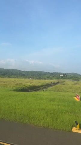 The video shows a wide, grassy field under a bright blue sky with scattered clouds. In the foreground, a dark asphalt road runs horizontally across the bottom of the frame. The field itself is predominantly a vibrant green, with patches of dried, yellowed grass interspersed. A narrow, shallow body of water, possibly a ditch or a small stream, cuts diagonally through the middle of the field. In the distance, a line of green, tree-covered hills forms the horizon, with a few white buildings visible nestled among the trees. The overall impression is one of a rural or undeveloped landscape on a clear day. There are no people or animals visible in the video.